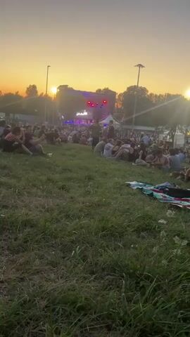A wide shot of an outdoor concert at sunset. A large crowd of people are seated on a grassy hill facing a stage in the distance. The sun is setting behind the stage, casting a warm orange glow across the sky. Lights are visible on the stage, and it appears to be a band performing. The people on the hill are casually dressed, and many are sitting on blankets or directly on the grass. The overall atmosphere seems relaxed and festive.