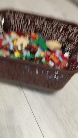 A brown woven plastic basket filled with small colorful toy building blocks is situated on a light gray wooden floor. The basket is in the center of the frame, and the camera pans around it. There is a dark gray blanket visible in the background on the right side of the image, and a white door on the left side.