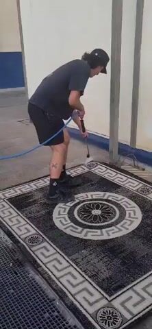 A person wearing a black t-shirt, black shorts, black socks, and black sandals is pressure washing a dark-colored rug with a grey and white geometric pattern. The person is holding a blue hose and a pressure washer wand. The rug is on a concrete surface. The person is bent over, directing the spray from the wand onto the rug. There is a white wall in the background.