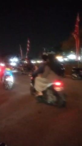 This is a blurry, dark video taken at night. The video shows a street scene with several motorcycles parked and one motorcycle moving from left to right across the frame, its taillight illuminated. Red and white flags are visible on poles in the background, and bright lights suggest a gathering or event. Several figures are present in the background, some appearing to be sitting. The overall impression is of a public space at night, possibly during a festival or market.