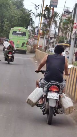 A person wearing a black sleeveless shirt and patterned shorts is riding a motorcycle on a paved road. The motorcycle has two large white containers strapped to its sides, one on each side, extending outwards. The motorcycle's license plate reads MH 25 Y 5999. The rider is facing away from the camera, looking towards the road ahead. In the background, a green bus is visible, along with other vehicles and buildings. The scene appears to be in an urban or semi-urban environment with trees and power lines present. The lighting suggests it is daytime.