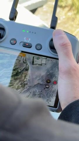 The video shows a close-up view of a person's hands holding a gray drone remote control. The remote has a screen displaying a map or aerial view of a rugged, mountainous terrain with some structures visible. The person's thumbs are positioned over the joysticks. The background is blurred, showing green foliage and a distant mountainous landscape under a partly cloudy sky. The video appears to be shot from the perspective of someone standing behind the person operating the drone.
