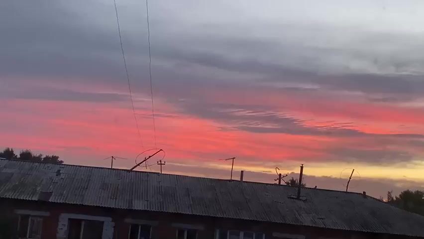 The video shows a building with a corrugated metal roof, several antennas, and a tall water tower in the background. The sky above is filled with dramatic clouds, painted in vibrant shades of pink, red, and yellow, indicating a sunset or sunrise. The light from the sky illuminates the scene from behind the building. Trees can be seen on the right side of the frame. The camera appears to be stationary, capturing the scene from a slightly elevated perspective.