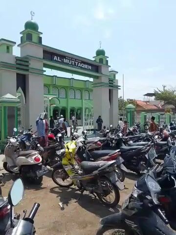 The video shows the entrance of a mosque named Masjid Jami Al-Muttaqien with a large parking lot filled with motorcycles. The mosque is painted in shades of green and white, and the entrance is a tall archway with the mosque's name displayed on top. People are seen walking in and out of the mosque. The parking lot is crowded with various models and colors of motorcycles. The sky is bright and clear. Some trees and buildings can be seen in the background beyond the parking lot.