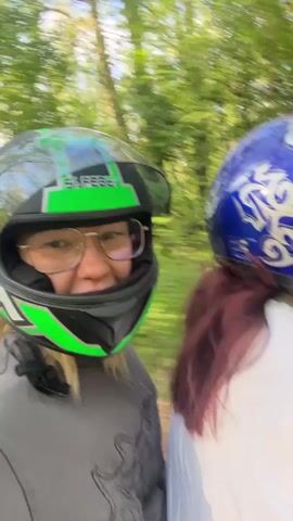 A video shows two people riding an ATV through a wooded area. Both riders are wearing helmets; one is green and black, and the other is blue with a white design. The person wearing the green helmet is filming, as they are looking directly into the camera. The ATV is traveling on a dirt path surrounded by trees. The weather appears sunny.