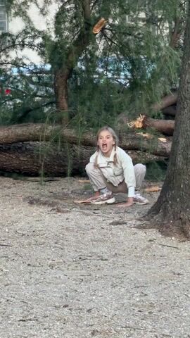 A young girl with blonde hair in pigtails crouches down outdoors on a gravel surface. She wears a white jacket, light-colored pants, and sneakers. She has her mouth open as if yelling. Behind her is a fallen tree trunk. She puts her hands down and moves like a crab, walking forward on her hands and feet.