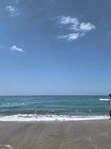 A lone person walks along a sandy beach next to a calm ocean under a blue sky with scattered clouds. The person is wearing a long-sleeved shirt and patterned pants and walks from right to left in the frame, with waves gently lapping at the shore.