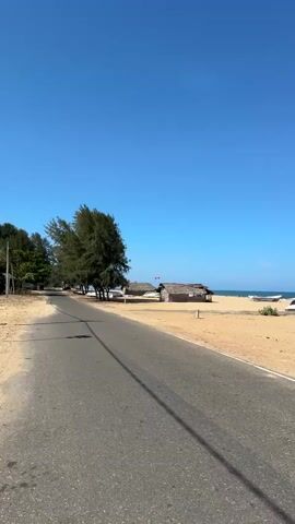 A short clip showing a paved road with a sandy beach to the right. On the beach are two thatched-roof huts and several boats. A clear blue sky is visible above. Two red flags are flying from one of the huts.