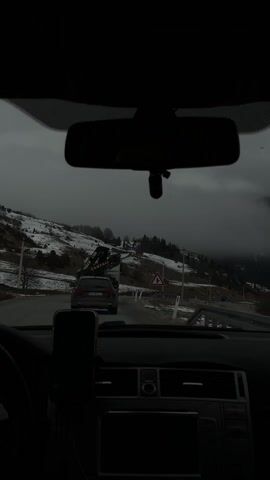 The video shows a car driving on a road in a mountainous area. The scenery is bleak, with snow-covered hills and cloudy skies. The road is winding and there is a truck in front of the car. There is a road sign indicating a yield to oncoming traffic. The interior of the car is visible, including the dashboard and rearview mirror.