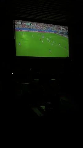 The video shows a large projection screen displaying a soccer match. The scoreboard indicates the teams are France and Spain, with the score at 0-0 in the 27th minute. The field is green, and many players in white and blue uniforms are visible. The projection is taking place in a dark environment, suggesting it might be nighttime or an indoor venue. Silhouettes of people watching the game can be seen in the foreground, obscured by the darkness. The overall impression is of a viewing party or a public screening of a soccer game.