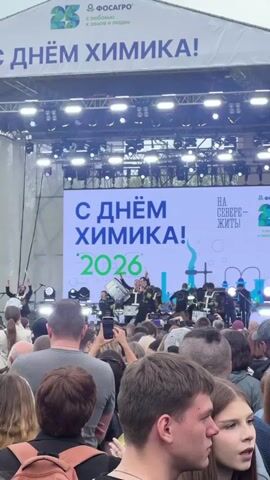 The video shows a crowd of people facing a stage decorated with the banners of “Fosagro” and the slogan “Happy Chemist’s Day!” in Russian. A band in black uniforms with gold detailing performs on the stage, while people in the audience record the performance with their phones. The stage features a large LED screen showing the text “Happy Chemist’s Day! 2026” and a logo with the text “To live in the north”. There are lighting fixtures above the stage. The crowd is diverse, consisting of people of different ages and genders. The environment suggests an outdoor event, possibly a festival or a company celebration.