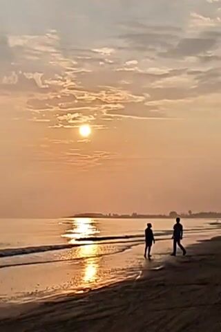 The video shows a beach scene during sunrise or sunset. The sun is visible in the sky, partially obscured by thin, patchy clouds. Two people are walking along the shoreline, their figures appearing as dark silhouettes against the bright light. The water reflects the sunlight, creating a shimmering path of light on the surface. The beach is sandy, and some tire tracks are visible on the sand. In the distance, there is a hint of land with some trees or buildings. The overall color palette is warm, with shades of orange, yellow, and gray dominating the scene.
