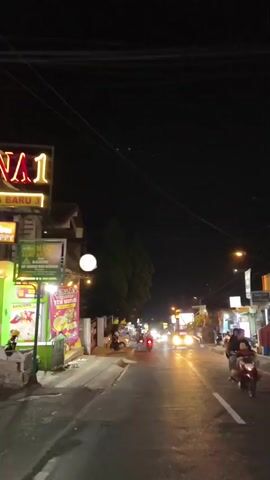 A night scene on a street with several shops and stalls lining the right side of the road. Cars and motorcycles are present on the road, some of which are in motion. The street is illuminated by streetlights and the lights from the shops. The sky is dark, indicating it is nighttime.