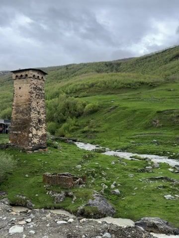 A stone tower stands on a green hill beside a flowing river under a cloudy sky. The tower is old and weathered, with narrow windows. In the background, more stone buildings are visible, as well as a lush green mountainside covered in trees. The river runs through the foreground, its water appearing murky. There are also some scattered rocks and debris along the riverbank. The overall impression is one of a remote and ancient settlement.