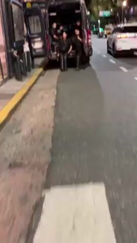 The video shows a nighttime urban scene with a street lined with buildings and trees. Several vehicles are visible, including police vans with officers standing near them. The officers appear to be in uniform, and some are getting out of or standing near the vans. Other cars are driving on the road. The street is marked with lane dividers. Streetlights illuminate the area. The camera viewpoint appears to be from someone standing on the sidewalk.
