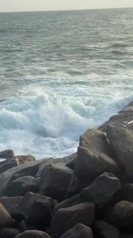 The video shows a rocky shoreline with waves crashing against the rocks. The ocean is a greenish-blue color, and the sky is a hazy yellow with some clouds. In the background, a lush green hill with palm trees can be seen. The sun appears to be setting or rising, casting a warm glow over the scene. The waves are white and foamy as they break against the shore. The rocks are dark and wet from the water. There are no people visible in the video.