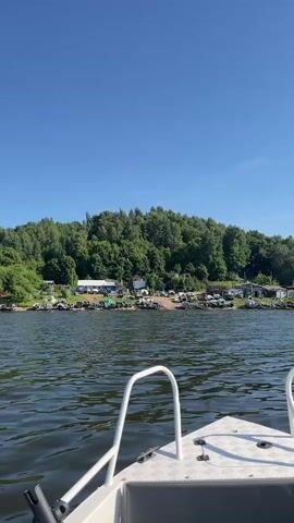This video depicts a serene day on the water in Siukterka, Russia. The perspective is from the bow of a small, white motorboat, moving gently across a dark, rippling body of water. The sky above is a clear, bright blue, indicative of a sunny day with good weather.

Along the distant shore, a line of trees forms a verdant backdrop. Among the trees, several small buildings or structures are visible, suggesting a settlement or recreational area. A number of vehicles, including cars and possibly trailers, are parked along the shoreline, hinting at an active day for visitors or residents. There are also some boats pulled up onto the bank.

The activity is the peaceful cruising of the motorboat on the water, likely for leisure or recreation. The overall mood is tranquil and inviting, capturing the essence of a pleasant summer day. There is no visible text within the video.