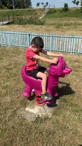 A young boy in a red t-shirt and black shorts is sitting on a pink spring rider shaped like an animal, possibly a dragon or a horse, in an outdoor park. The boy is holding onto the handlebars of the spring rider with both hands and appears to be bouncing. He is wearing blue and yellow sandals and yellow socks. The spring rider is made of plastic and has red springs underneath. The boy is positioned on a patch of dry grass. In the background, there is a light blue and white wooden fence. Beyond the fence, there is a grassy hill with some trees and a set of stairs leading up the hill. The sky is bright and appears to be a sunny day.
