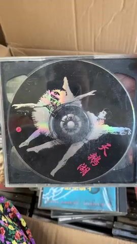The video shows a cardboard box filled with CDs. In the center of the box, a CD case is open and displaying the CD itself. The CD has an image of two figures, possibly dancers, in white costumes against a black background. There are some Asian characters written on the CD. To the left of this CD is another CD case with a label that reads "NATURE'S BEAUTY" and shows an image of a horse-drawn carriage. To the right of the central CD is another CD case labeled "TCHAIKOVSKY MASTERPIECES" featuring a portrait of a man. Below the central CD, there are more CD cases, one of which has a label that includes "SYMPHONIE NO. 6 PATHÉTIQUE." The overall impression is that of a collection of classical music CDs stored in a box.