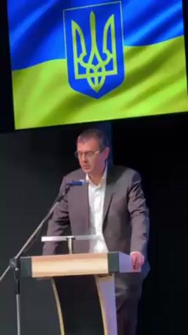 A man in a brown suit and white shirt is speaking at a podium. He is wearing glasses and looking to his left as he speaks. Behind him is a large screen displaying the flag of Ukraine, with the Ukrainian coat of arms prominently displayed. The podium is made of light wood. The background behind the screen is black.