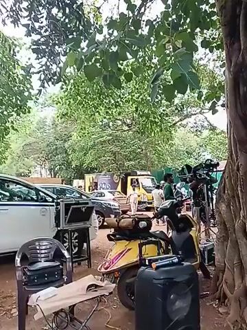 The video shows a behind-the-scenes view of what appears to be a film or video shoot taking place outdoors, possibly in a park or wooded area. There are trees overhead with dense foliage, creating a canopy.  Several vehicles are parked, including a white van and a yellow service truck with what looks like a logo on the side, as well as at least two other cars. A yellow scooter is parked in the foreground, close to camera equipment, a small table, and a plastic chair. A group of people are standing in the background, some appearing to be crew members around video cameras and equipment. The setting suggests a production set, with various elements in place for filming.