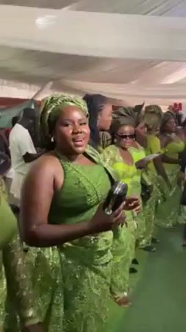 The video shows a group of women dressed in coordinated green outfits, likely at a wedding or similar event. They are standing in a line, possibly waiting to participate in a procession. The women are wearing matching headwraps and dresses, some with lace details. The event appears to be taking place under a large white tent. Some of the women are holding programs or leaflets. The environment seems lively and festive.