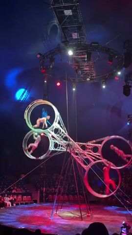 A circus act features performers inside large, spinning metal rings. The rings are part of a tall, multi-tiered structure. The top of the structure is illuminated by spotlights, and the background is a dark blue circus tent ceiling. Several performers are visible within the rings, some appearing to be suspended or moving in coordination with the spinning. The lighting is dramatic, with blue and red hues illuminating the stage and the performers. An audience is seated in the background, observing the performance.