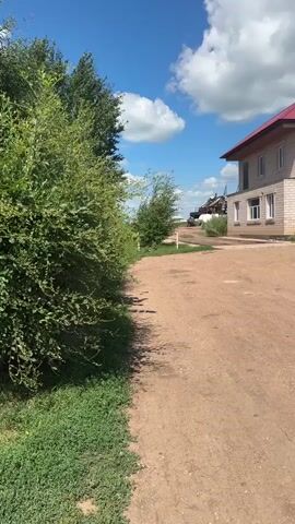 The video shows a short clip of a dirt path next to a dense green hedge. On the right side of the path, there is a building that appears to be a light-colored, two-story structure with a red roof. In the distance, there are some other structures and trees. The sky is blue with some white clouds. The sun is shining, casting shadows on the path. The camera is moving slowly forward, showing the path and the hedge.