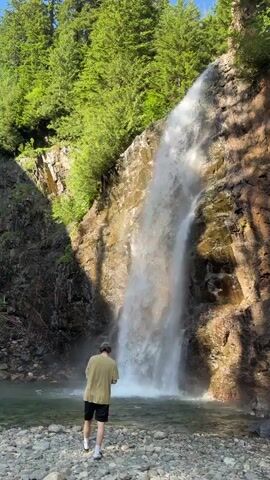 A person stands at the base of a waterfall, looking at a phone. The waterfall is cascading down a rocky cliff face surrounded by lush green trees. The ground in front of the waterfall is covered with grey pebbles and some water. The person is wearing a tan shirt and black shorts. The scene is illuminated by sunlight.