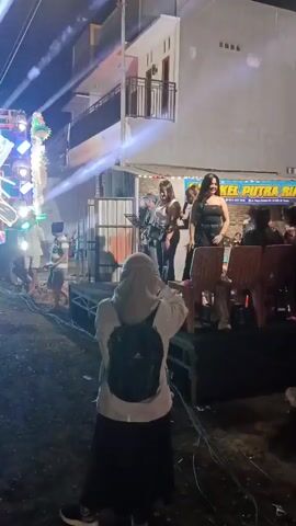 The video shows an outdoor nighttime performance with a small stage set up. There are at least three female performers on the stage, two of whom are singing or speaking into microphones. One performer in a black dress is dancing and gesturing. In the foreground, a person wearing a headscarf and a backpack is filming the performance with their phone. There are lights and sound equipment set up around the stage, and an audience is visible sitting and standing in the background. A building is visible behind the stage, with a sign that reads "BETEL PUTRA RIAN". The ground is covered in dirt or gravel.