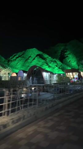 The video shows a nighttime view of a park or resort area with buildings and mountains illuminated in green light. The camera moves slowly along a paved path. The buildings appear to be restaurants or recreational facilities. A small playground is visible in the background. The mountains form a backdrop, brightly lit with green lights to emphasize the vegetation. The area seems designed for leisure and relaxation, with the lighting creating a visually striking scene.