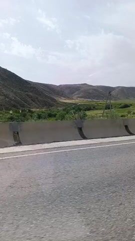 The video shows a view from a moving vehicle on a paved road. A concrete barrier runs along the side of the road, separating it from a grassy area and hills in the background. The hills are covered in sparse vegetation, and the sky is cloudy. The time stamp indicates the video is a short clip, likely from a dashcam or phone recording. There are no people or other vehicles visible in the frame.