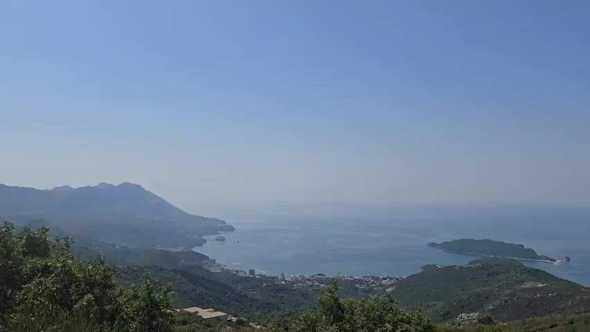 A panoramic view from a high vantage point shows a vast, calm blue sea under a clear, pale blue sky. In the distance, a coastline is visible with green, rolling hills and mountains. Along the coast, a town or city with white buildings and some taller structures is spread out. A small island is situated in the sea off the coast, connected to the mainland by a narrow strip of land or a bridge. The foreground is dominated by lush green trees and vegetation. The overall scene depicts a scenic coastal landscape on a clear day.