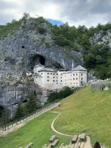 This video pans across a scenic view of a cliffside castle. The castle is built into a large cave opening in a rocky mountain face. The castle is white with multiple windows and a few towers. There is a pathway leading up to the castle with a wooden railing. The area in front of the castle is a grassy hillside with some wooden benches and picnic tables. In the background, there are trees and lush green vegetation covering the mountainside. The sky is partly cloudy.