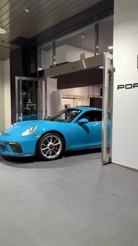 A bright blue Porsche 911 GT3 is captured slowly driving out of a Porsche dealership or showroom. The vehicle is positioned between two large, automatic glass doors that are opening to facilitate its exit. A person is visible in the driver's seat.

To the right of the car, a prominent white wall displays the "PORSCHE" text in black capital letters, with the brand's crest logo positioned above it. The interior of the showroom, featuring a tiled floor and overhead lighting, is visible through the glass doors in the background. The car has a Japanese license plate on its front bumper, partially readable as "... 99-99". The setting is an indoor environment, likely a modern dealership in Minato, Japan.