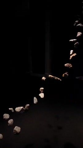This is a vertical, low-angle video showing a dark environment. A scattering of rocks is arranged in a curved line, starting from the bottom left and curving upwards towards the right. The rocks are illuminated by a beam of light from above, casting shadows on the dark surface below. The rocks appear to be suspended or floating, although this may be an illusion created by the lighting and arrangement. There are no people or other visible objects in the scene. The overall mood is mysterious and somewhat unsettling due to the darkness and the unusual presentation of the rocks.