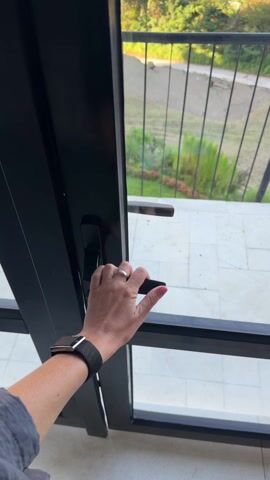 A first-person perspective video shows a hand with a ring and a smart watch opening a glass door leading to a balcony. The balcony has a metal railing. The view from the balcony is a landscape with a large mountain in the distance, surrounded by green vegetation and a few scattered buildings. A road runs through the lower part of the scene. The sky is blue with a few clouds. The person opens the door and pauses, taking in the view.