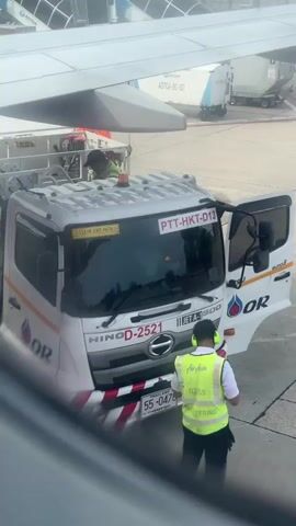 A person in a yellow vest and ear protection stands next to a white truck with "HINO" and "D-2521" written on it. The truck has "PTT-HKT-D13" on its windshield. The person holds a clipboard and appears to be inspecting the truck, which has an "OR" logo on its side. In the background, an airplane wing is visible, and the tarmac is in view.