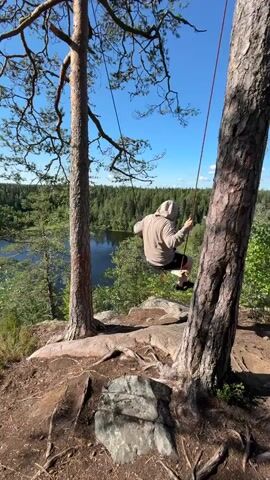 The video captures a person on a swing set on a cliff overlooking a forest and a lake. The person is wearing a hooded sweatshirt and shorts, and is holding onto the swing ropes. The swing is attached to a tree. The sky is blue and clear, and the sun is shining. The trees are green and lush, and the lake is calm and still. The overall impression is one of peace and tranquility.