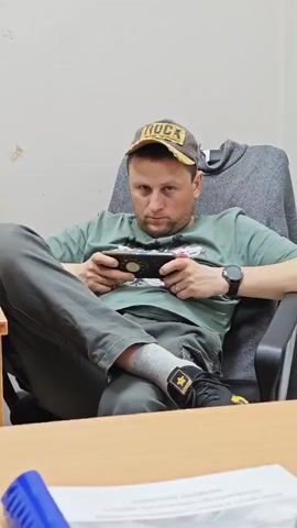 The video shows a man sitting in an office chair behind a desk, playing a game on a handheld console. The man is wearing a green t-shirt, gray pants, socks, and black shoes with gold stars. He also has a watch on his left wrist and a baseball cap that says "ROCK" on the front. The chair is black and gray. The desk is light brown. There is a gray hoodie draped over the back of the chair. There are some papers and a blue pen on the desk. The wall behind him is white. He looks up and smiles at the camera at one point.