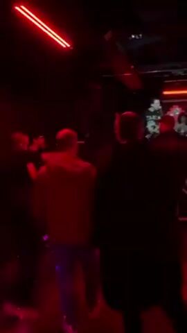 The video captures a live music performance in a dimly lit venue bathed in red light. A band is performing on a raised stage with multiple vertical and horizontal red lights surrounding them. The band members include a singer, guitarists, and a drummer, all silhouetted against the stage lights. A crowd of people stands in front of the stage, mostly visible as dark shapes. Some audience members are moving, possibly dancing or interacting with each other, while others appear to be watching the performance. The overall atmosphere is dark and energetic.