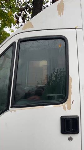 A short video starts with a view of the side of a white van. Raindrops are visible on the van window. Inside the van, an older, bald man is seated in the driver's seat. The camera zooms in on the man, and a hand places a small, cartoon sticker on his forehead. The camera focuses closely on the sticker. The man has gray hair on the sides of his head and blue eyes. He smiles slightly during the close up.