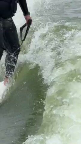 The video shows a person wake surfing. The individual is wearing a black wetsuit and holding onto a rope handle. They are standing barefoot on a white wakesurf board, riding the wake of a boat. The water is churning around the board, creating white foam. The day appears overcast.