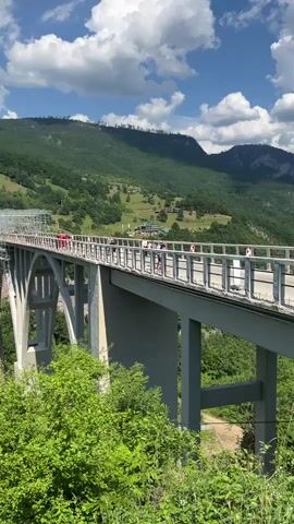 The video shows a wide shot of a large concrete bridge spanning a deep gorge. The bridge has an arched central section and is supported by massive concrete pillars. Scaffolding is visible on one side of the bridge, suggesting ongoing construction or maintenance. Several people are walking on the pedestrian walkway of the bridge. The gorge is covered in lush green trees, and the surrounding landscape consists of rolling hills and mountains also covered in dense forest. The sky is blue with scattered white clouds. The camera pans slowly to the right, revealing more of the gorge and the mountainous terrain.