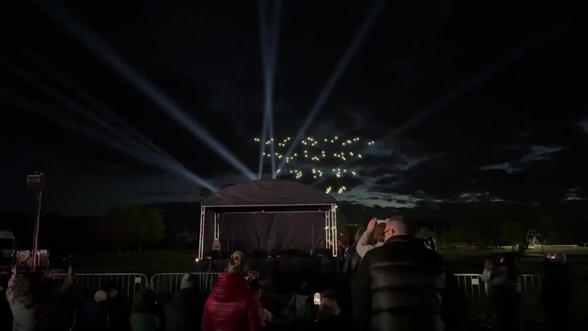 At night, a crowd is standing behind a metal fence, facing a stage with lighting on it. Above the stage are lights that look like drone lights, forming a display in the sky. People are looking at the stage and the sky, many holding up their phones to record. There are trees and green space in the background. The sky is dark, with some cloud cover. White light beams emanate upwards from behind the stage.