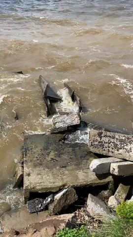 The video shows a body of murky brown water with waves crashing against broken concrete debris. The concrete is in various sizes and shapes, some pieces are large slabs, while others are smaller chunks. The debris is partially submerged in the water and appears to be part of a collapsed structure. The water is choppy and turbulent, suggesting a strong current or wind. There are no people or animals visible in the video. The scene is located at the edge of the water, with some dry land and green vegetation visible in the lower right corner of the frame. The overall impression is one of decay and destruction.