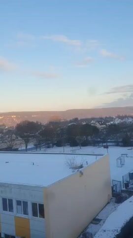 The video presents a high-angle view of a snow-covered schoolyard in a European-looking town. The sky is blue with light cloud cover.  The school building has a flat roof, also covered in snow, with white walls and yellow accents.  A French flag is visible on the building.  The schoolyard itself is covered in a layer of snow, with patches of cleared pathways and vegetation, including trees and bushes, also covered in snow.  A group of children and possibly some adults are present in the schoolyard, some near a fenced area.  In the distance, a residential area with snow-covered rooftops can be seen, with hills visible in the background.  The overall impression is of a cold, wintery day at a school in a small town.