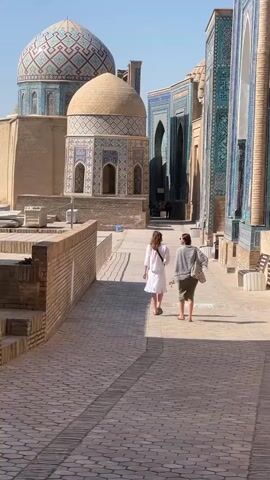 This media file showcases two individuals walking down a sun-drenched pathway paved with cobblestones. The pathway is flanked by low brick walls on one side and ornate, tile-covered buildings on the other. In the background, two prominent domes are visible: one larger, intricately patterned with geometric designs in blue, white, and brown; and a smaller, lighter brown dome adorned with similar decorative tiling and arched windows. To the right, a taller structure with blue and turquoise tilework, featuring arched doorways, stands prominently. The scene appears to be a historical or religious site, likely a complex of mausoleums or mosques, given the architectural style and the presence of domes and decorative tiling characteristic of Central Asian Islamic architecture.

The time of day appears to be morning or afternoon, indicated by the bright sunlight casting distinct shadows on the pathway. The sky is clear and blue, suggesting fair weather. The two subjects, viewed from behind, are dressed casually. The person on the left wears a white dress, while the person on the right is in a green skirt and a gray cardigan. They are walking away from the camera, towards the buildings in the distance. One of the individuals appears to be holding a phone, possibly taking pictures. The overall mood seems peaceful and contemplative, as the individuals explore the historic architecture under a bright sky. No discernible text is visible in the media file. The location context of Samarkand City, Uzbekistan, strongly suggests this is a view of the Shah-i-Zinda necropolis, famous for its stunning tilework and mausoleums.