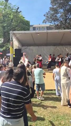 A group of people are performing traditional dances on a wooden stage outdoors. Several performers are wearing brightly colored, traditional clothing. Some are playing instruments like accordions. A crowd of spectators is gathered in front of the stage, watching the performance. The scene is set on a sunny day, with trees and buildings visible in the background. The image is a still frame from a video.