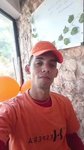 The video shows a man in an orange shirt and baseball cap sitting in front of a stone wall with a whiteboard. He smiles at the camera and then someone calls out "Nena." He is joined by two people, one lying down and one standing. The person lying down is also wearing an orange shirt. The person standing is wearing a black and white striped crop top. They are in a bright room with a window with the letters "HPA" visible. The standing person leans toward the camera and makes a peace sign. The person lying down has a pen in their hand and is looking at their phone.